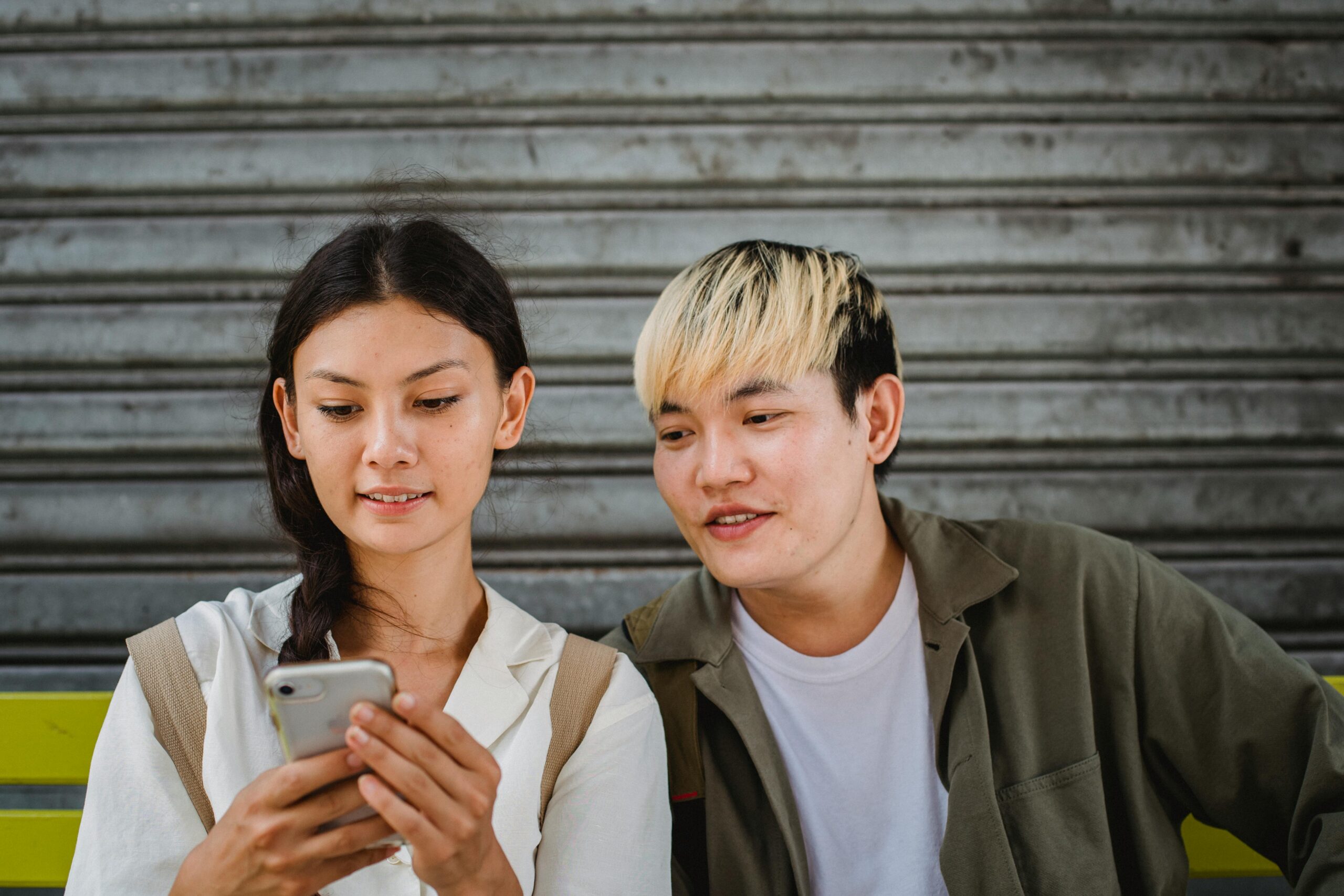 Content young Asian couple in casual outfits surfing contemporary mobile phones and sitting on street bench on clear weather