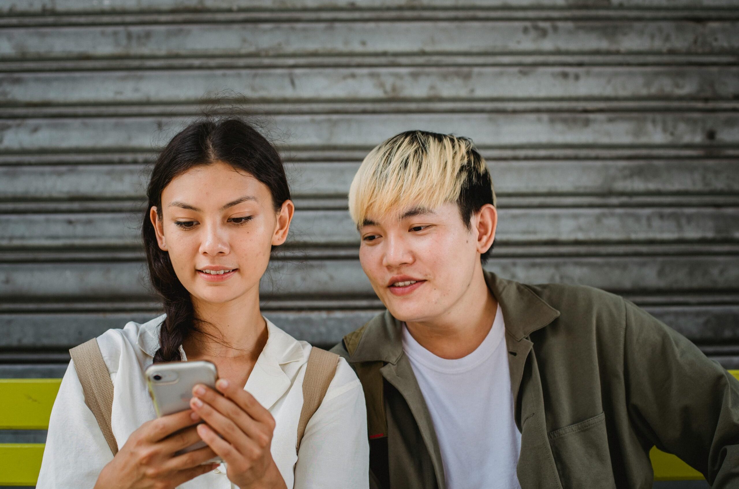 Content young Asian couple in casual outfits surfing contemporary mobile phones and sitting on street bench on clear weather